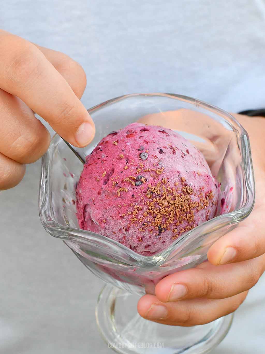 Child holdingmixed berry ice cream topping with chocolate shavings in a glass bowl